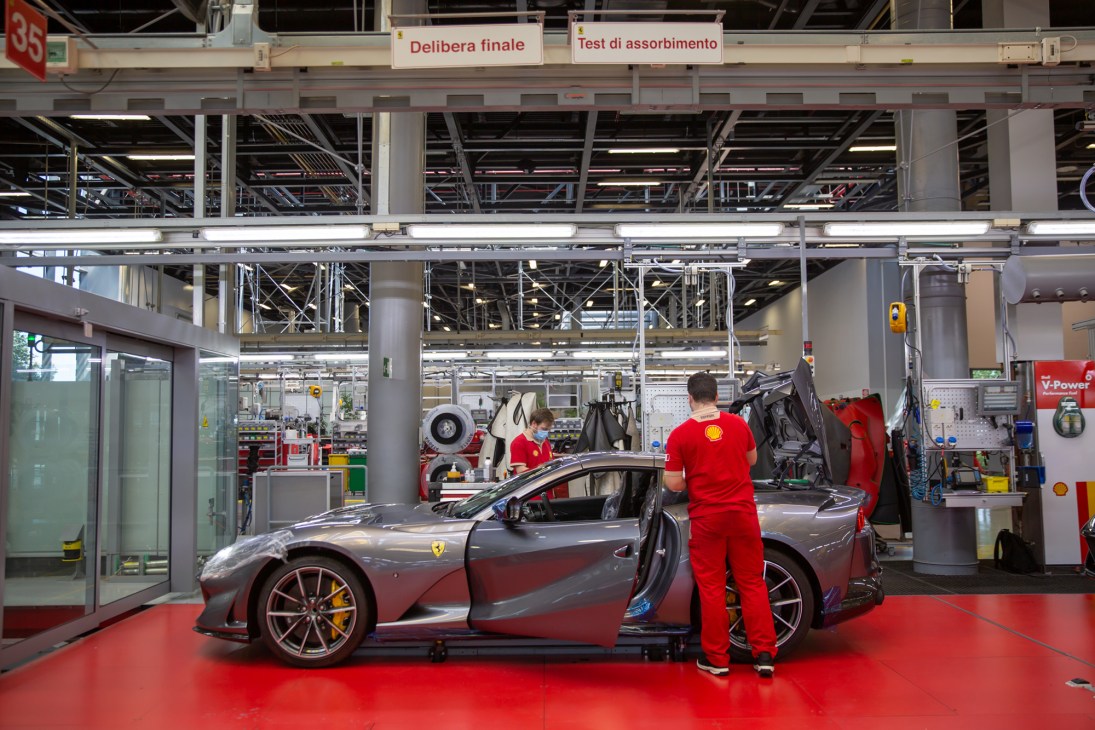 Ferrari technicians in red uniforms working on a gray sports car in a modern factory facility