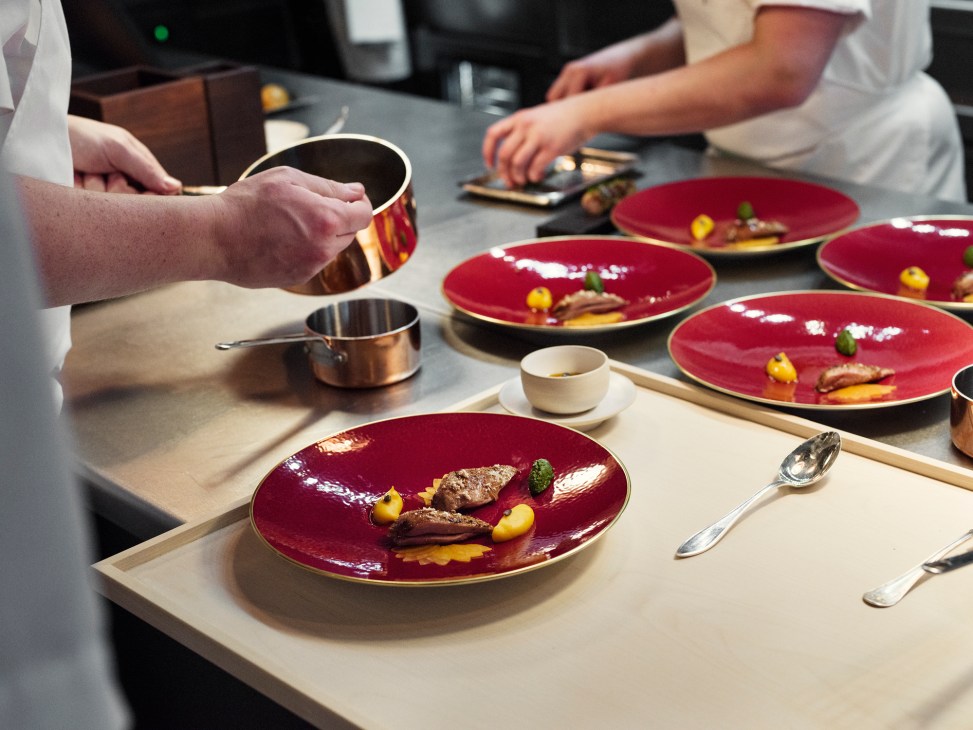 Chefs plating elegant dishes on red plates with gold trim in a professional kitchen