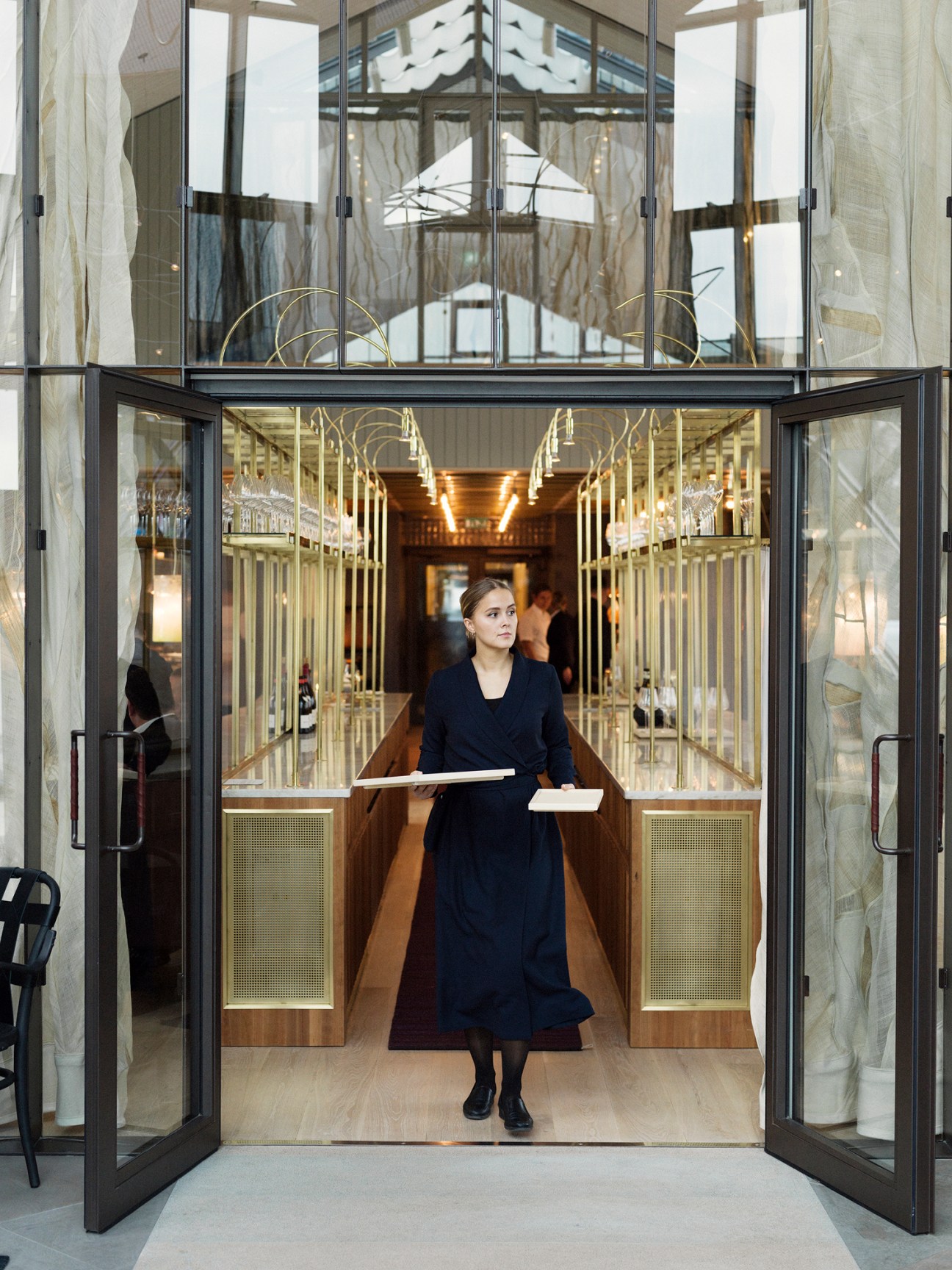 Woman in navy dress carrying plates through glass doors of restaurant service station with marble, brass and cherry wood