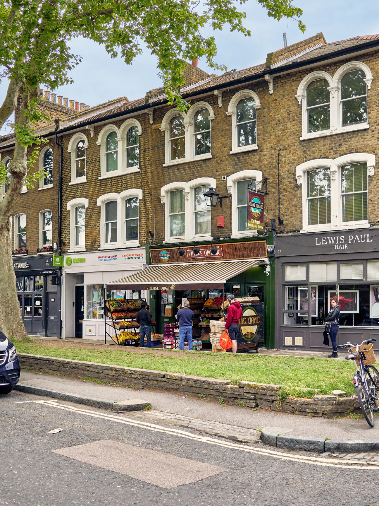A street-level view of small shops including Village Organic and Lewis Paul Hair on an East London street with Victorian buildings above.