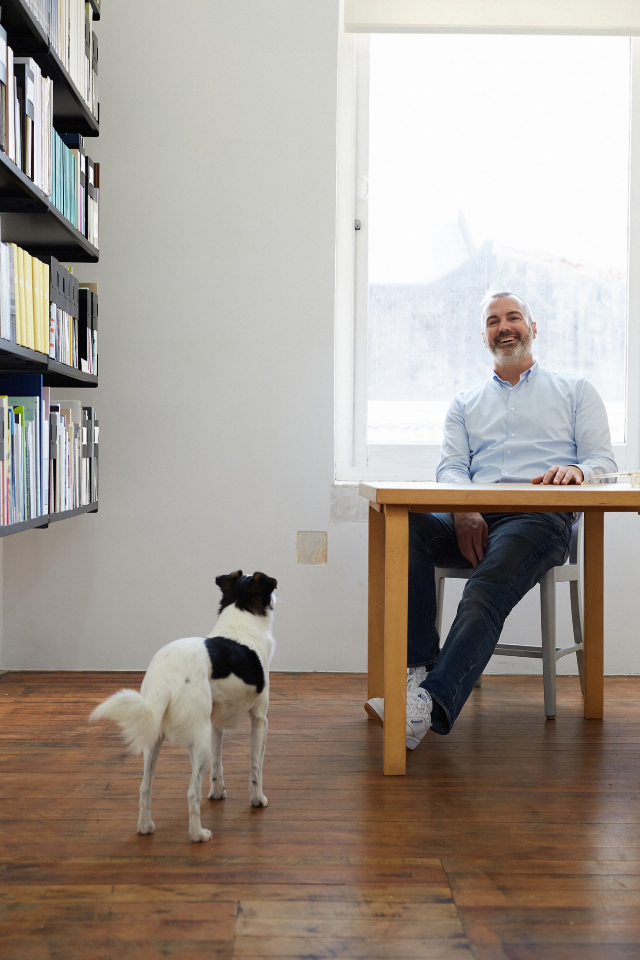 Man in light blue shirt sitting at wooden table by large window with black and white dog standing nearby.
