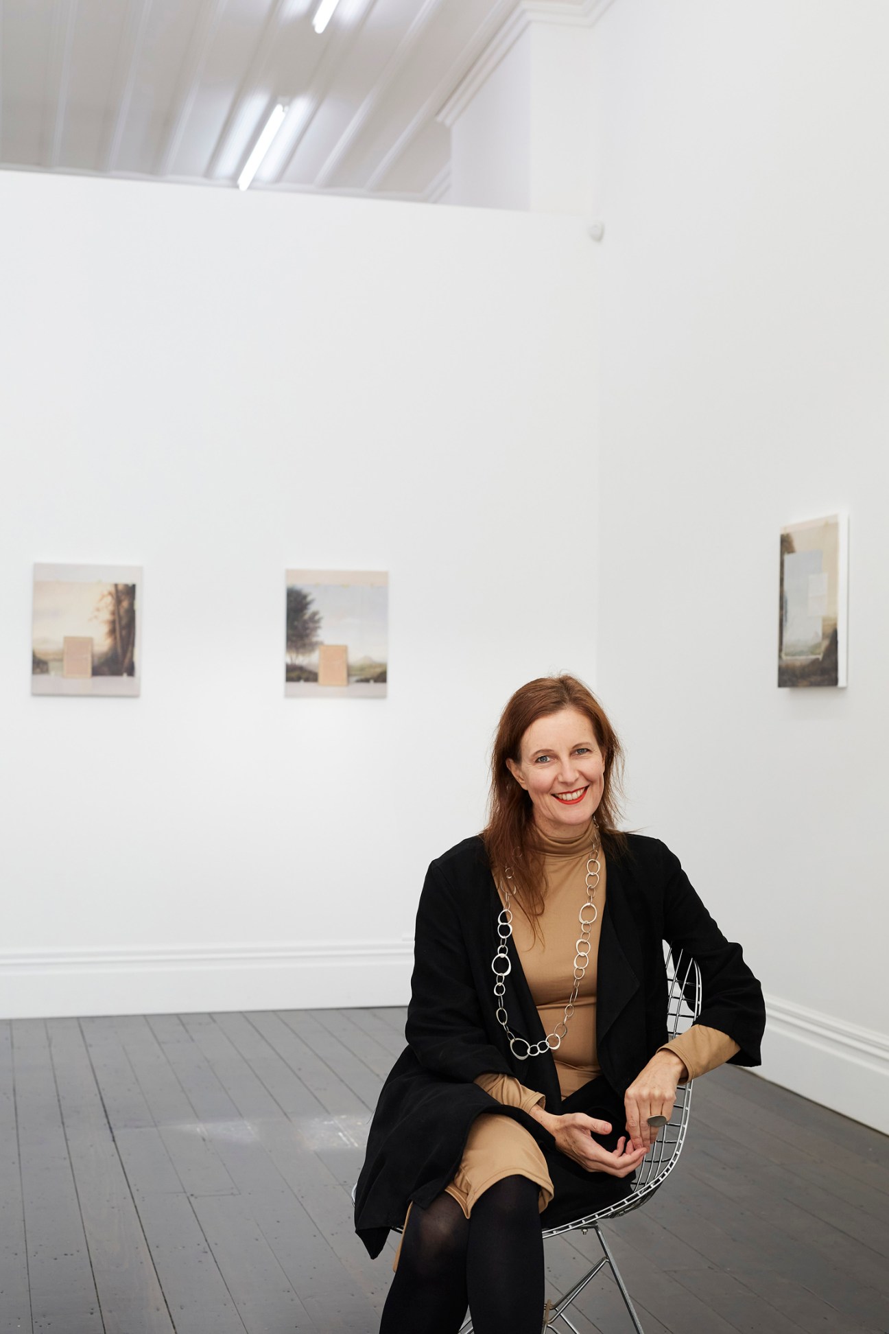 Melanie Roger, gallerist, sits in a white gallery space with small framed artworks on the wall behind her.