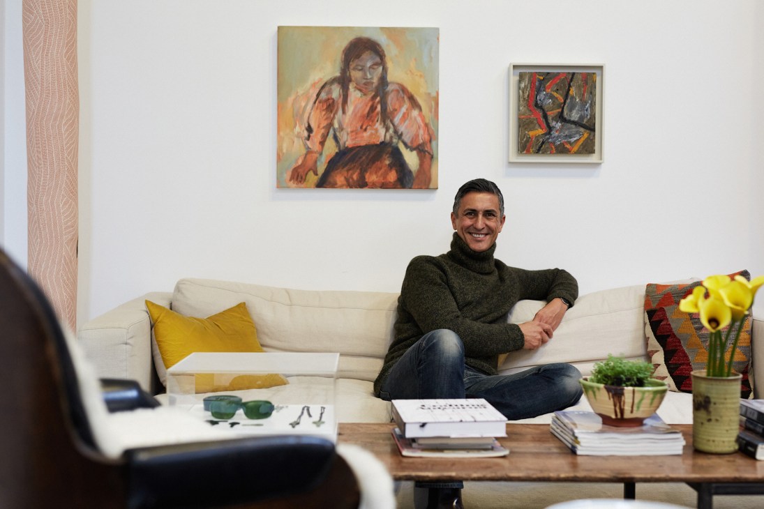 Tim Melville sits smiling on a white couch in his gallery space with artwork on the walls behind him