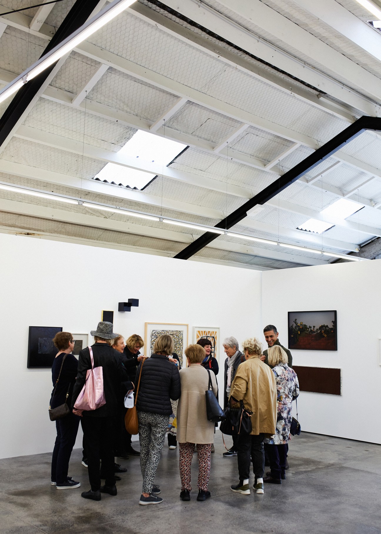 Group of gallery visitors examining artworks displayed on white walls in a modern industrial gallery space.