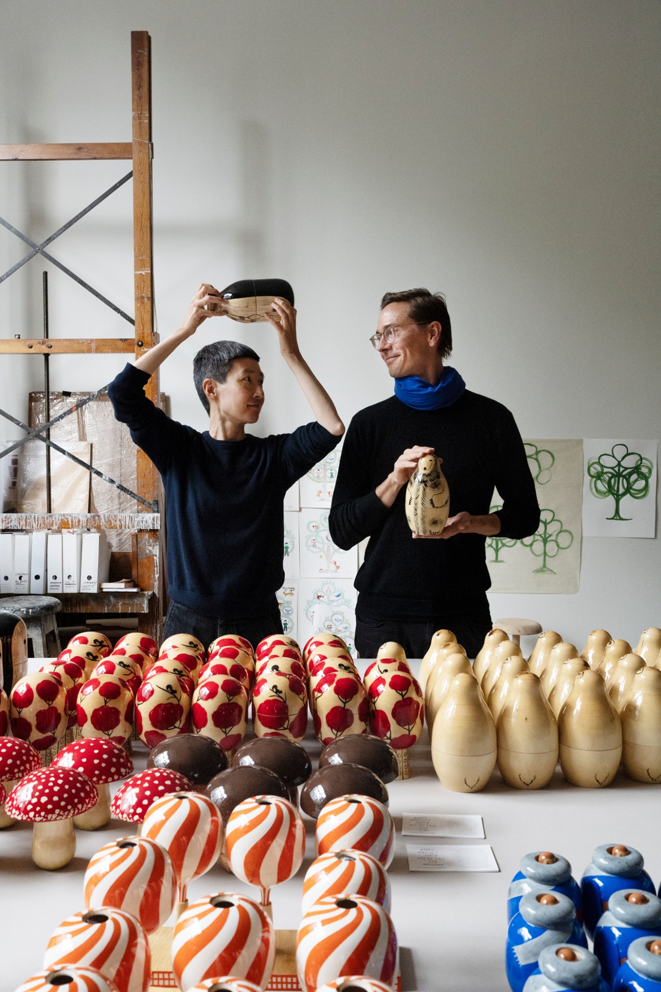 Aamu Song and Johan Olin examining wooden nesting dolls in their studio workspace filled with colorful handcrafted objects