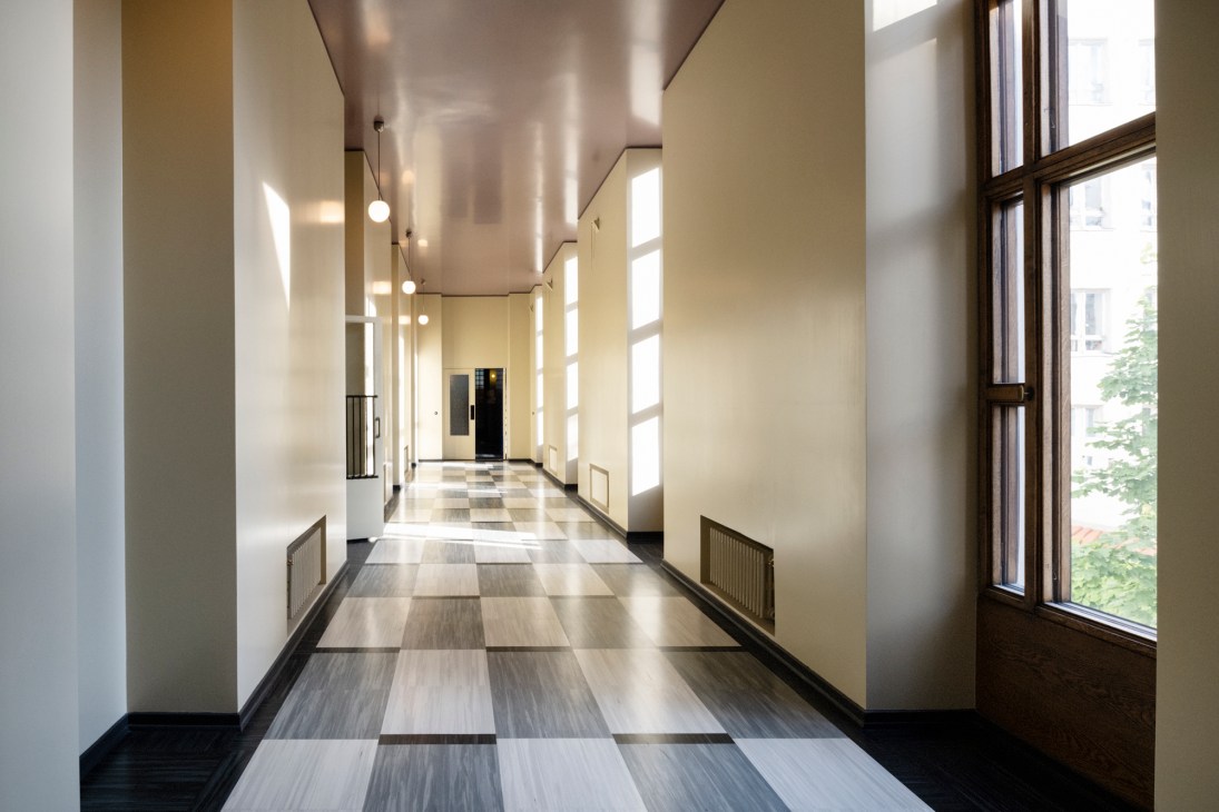 Long interior hallway with checkered marble flooring, tall windows, pendant lighting and cream-colored walls