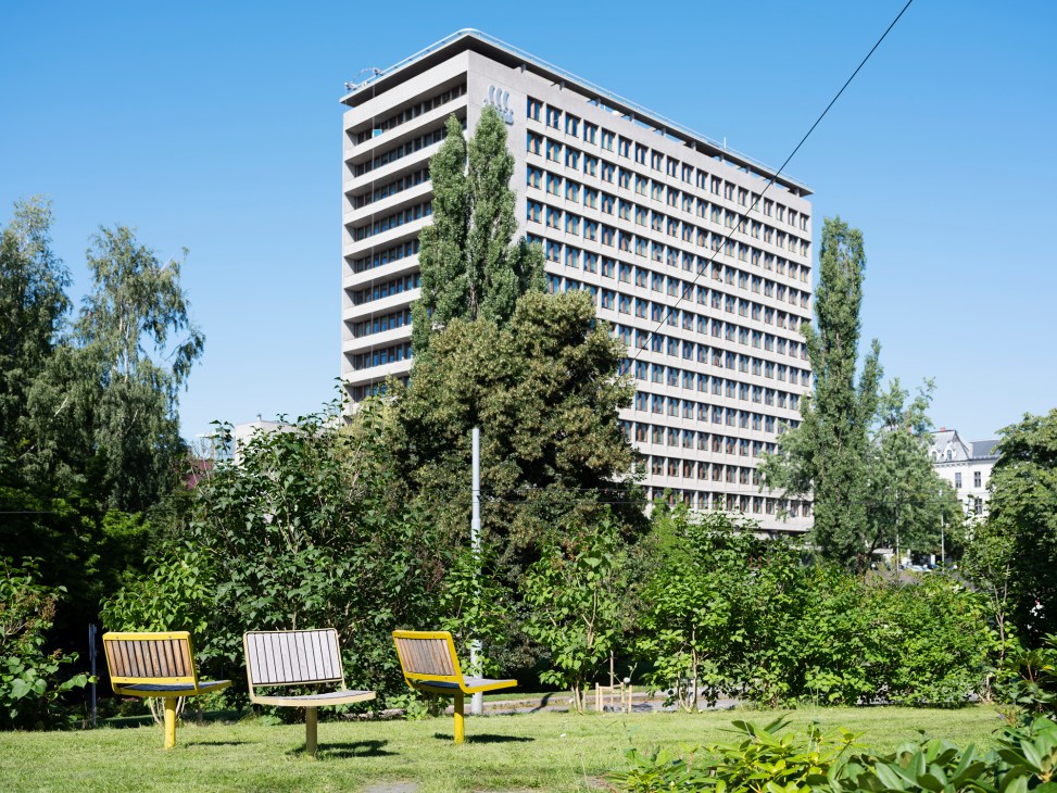 Modern multi-story residential building with rows of blue-framed windows surrounded by green vegetation and outdoor seating.