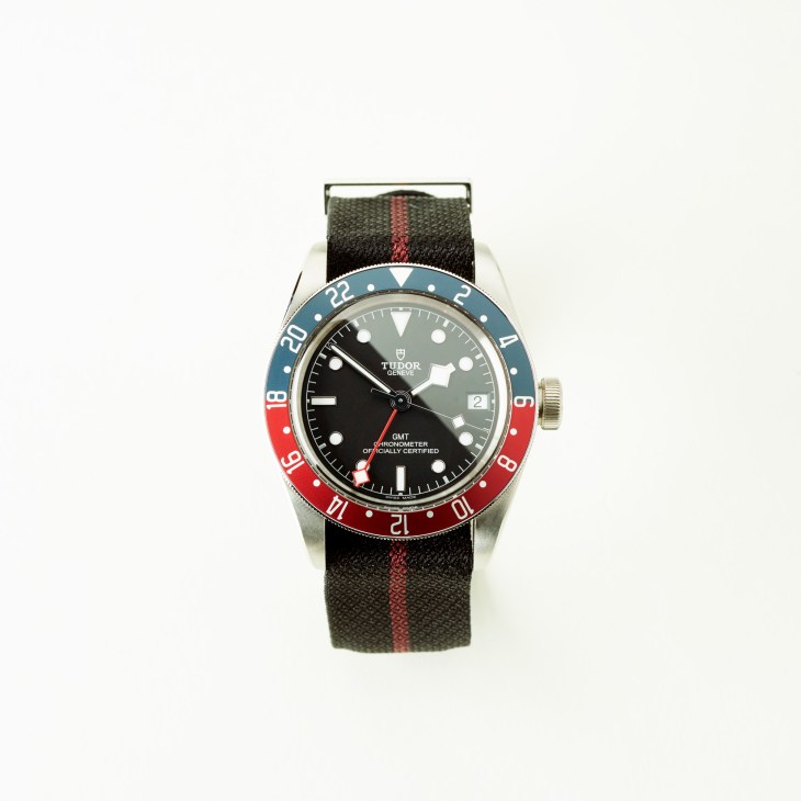 Tudor Black Bay GMT watch with black dial, blue and red bezel, and fabric strap with red stripe.