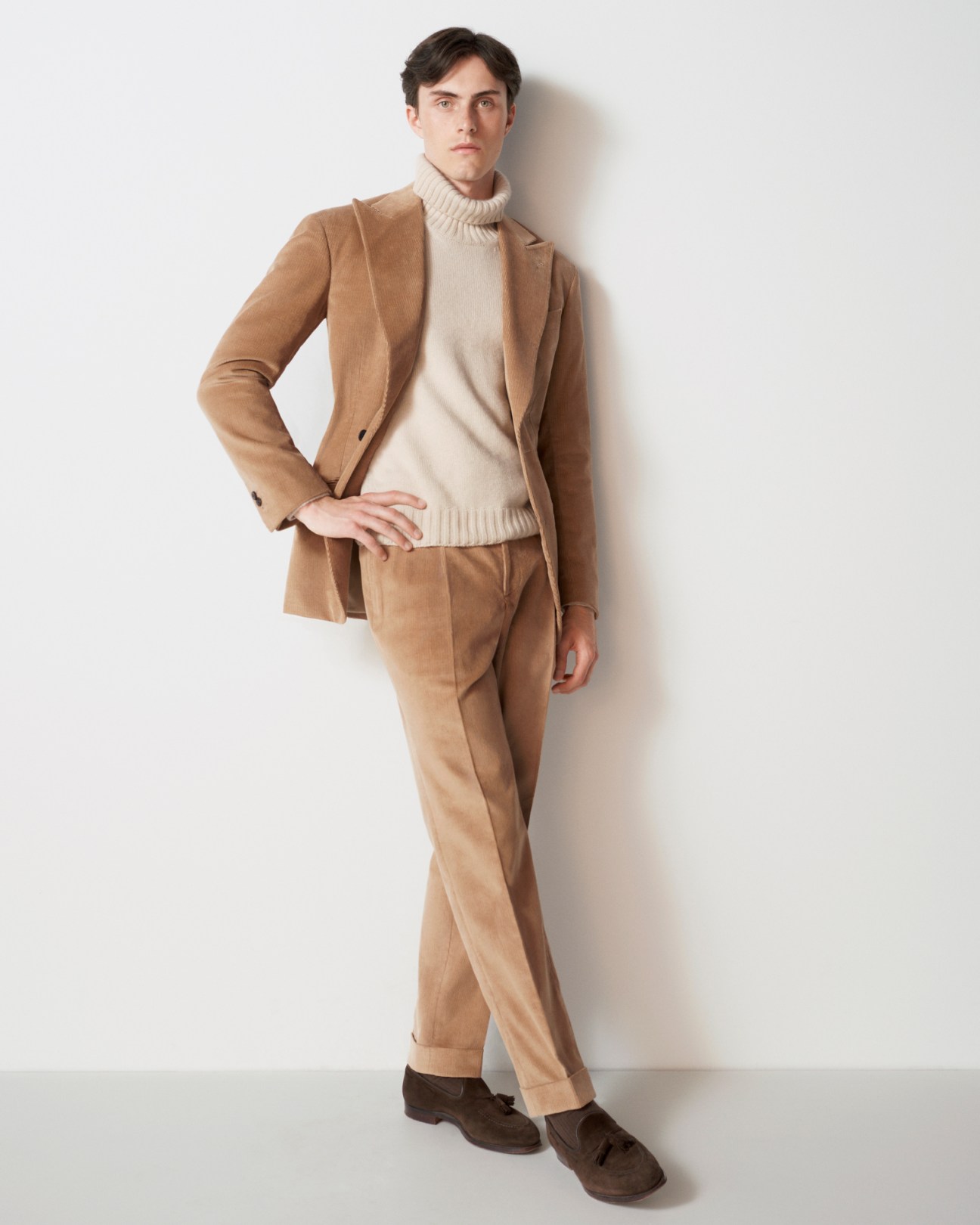 Man in tan corduroy suit with cream turtleneck sweater and brown suede shoes against white background