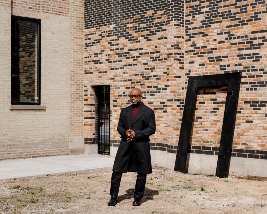 Theaster Gates standing in front of brick building wearing long black coat and glasses