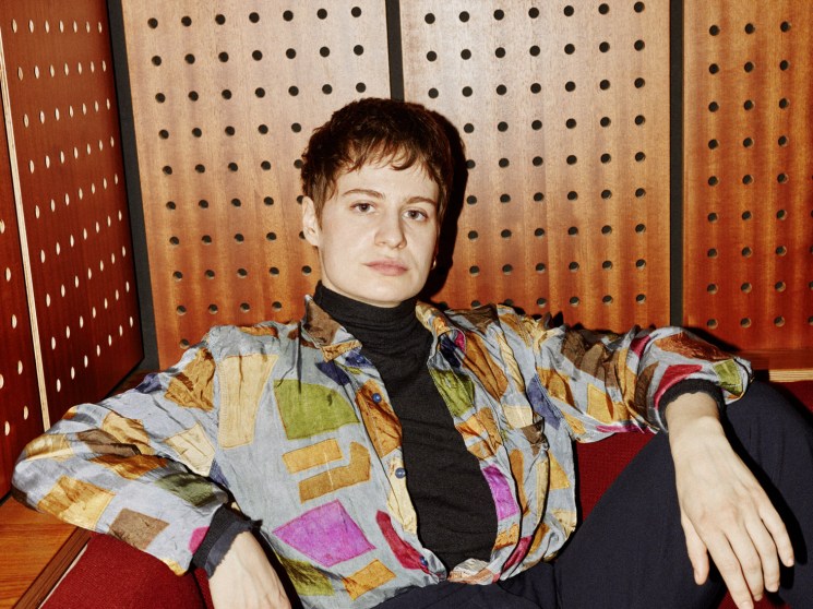 A short-haired person in a colourful printed shirt and black turtleneck reclines in a red chair against a warm wood-panelled wall.