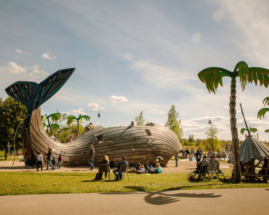 Wooden whale-shaped playground structure with children playing, surrounded by tropical plants and visitors in a park setting.