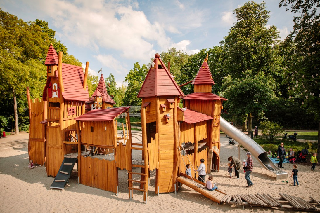 Wooden castle-themed playground with red-roofed turrets and a metal slide, surrounded by children and trees.
