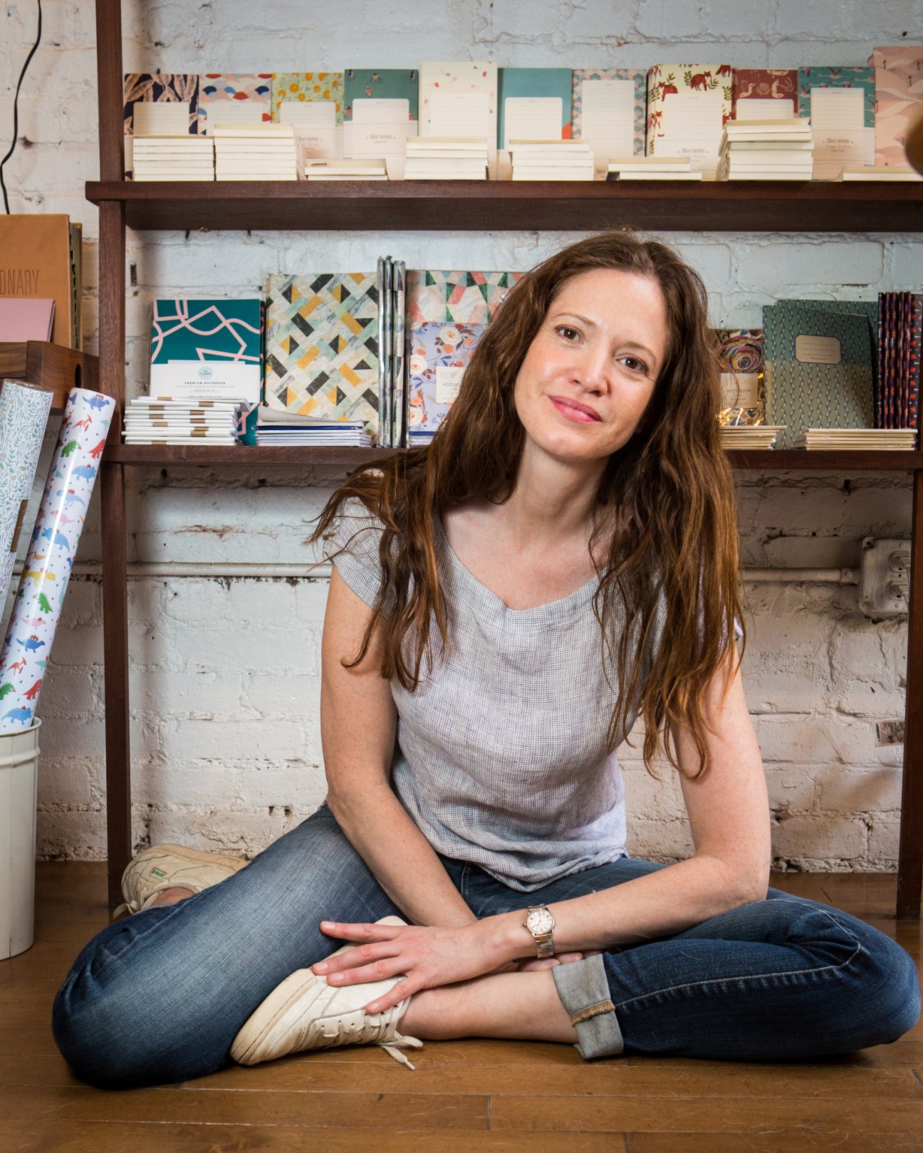 Woman with long brown hair sitting cross-legged in front of shelves displaying stationery and notebooks.