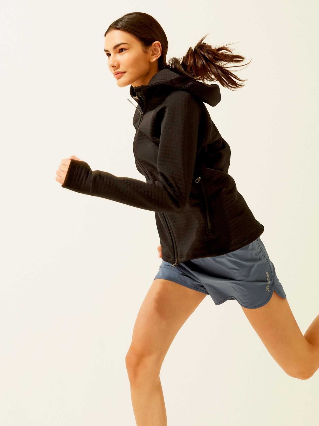 Woman jogging in black hooded jacket and blue shorts against neutral background