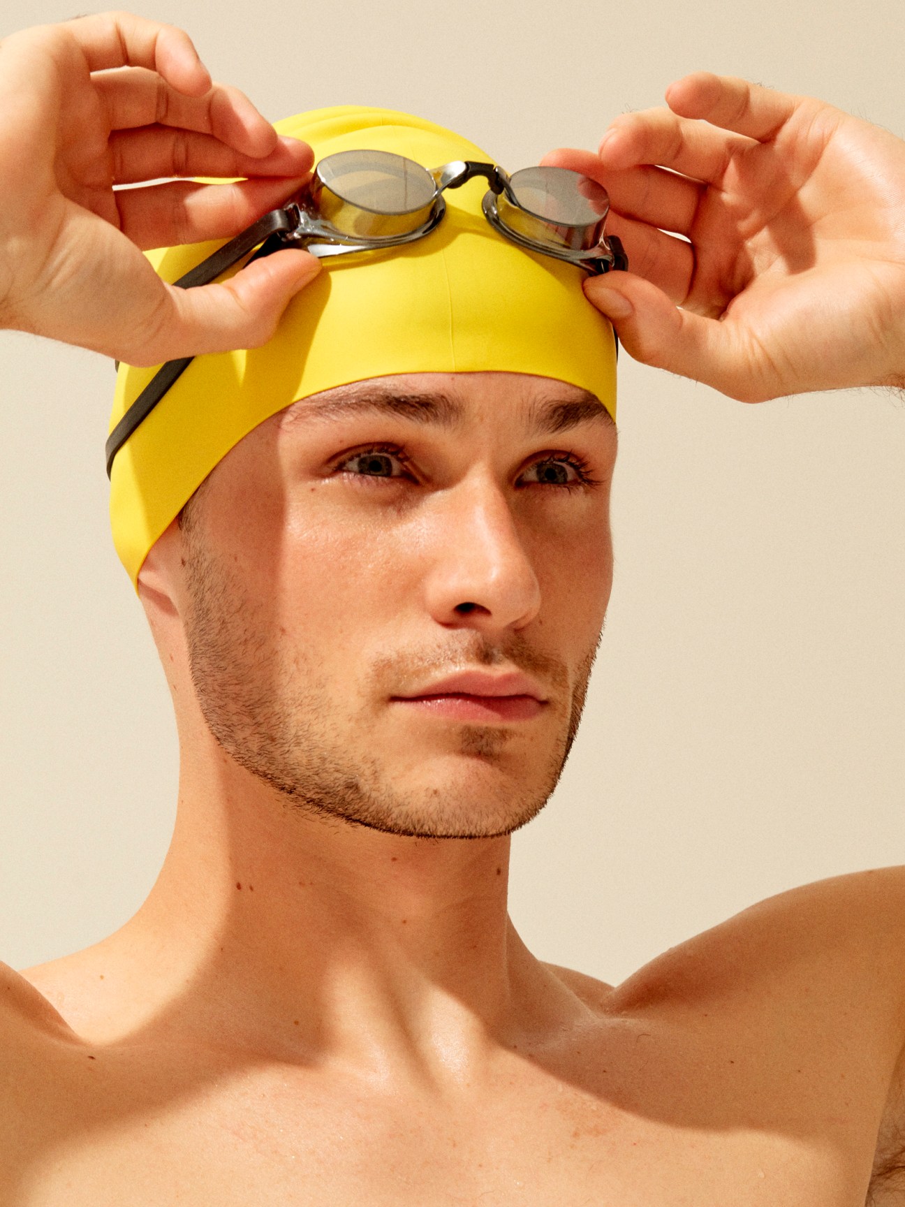 Man wearing a yellow swimming cap holding up mirrored swim goggles.