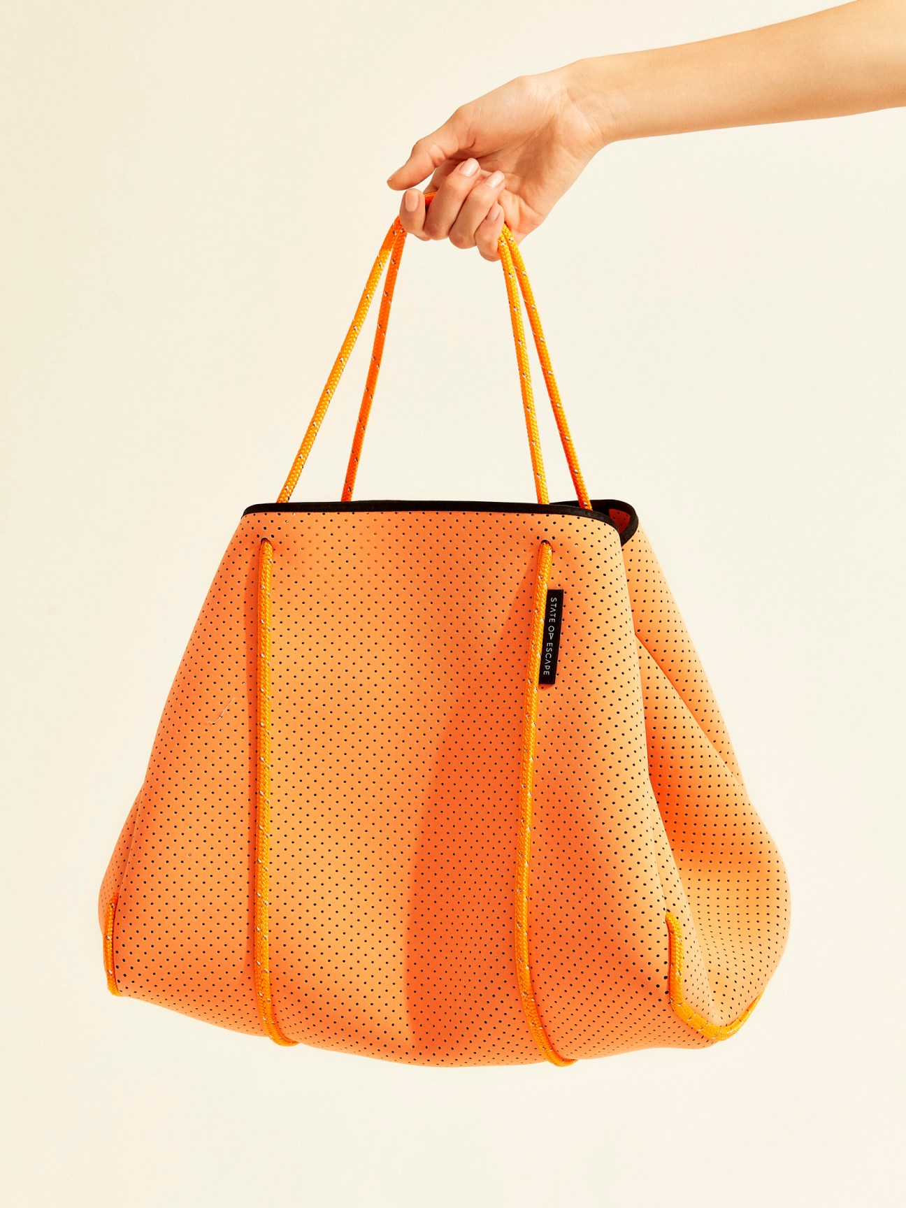 Hand holding an orange neoprene State of Escape tote bag with yellow accents against a cream background.