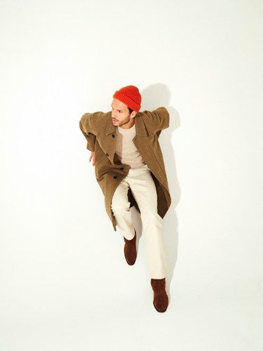 Man wearing olive-brown jacket, cream sweater, white trousers, red beanie, and brown boots against white background.