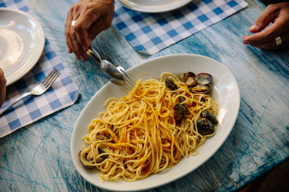 Hands twirling spaghetti with clams on a fork from a white oval plate on a blue checkered tablecloth