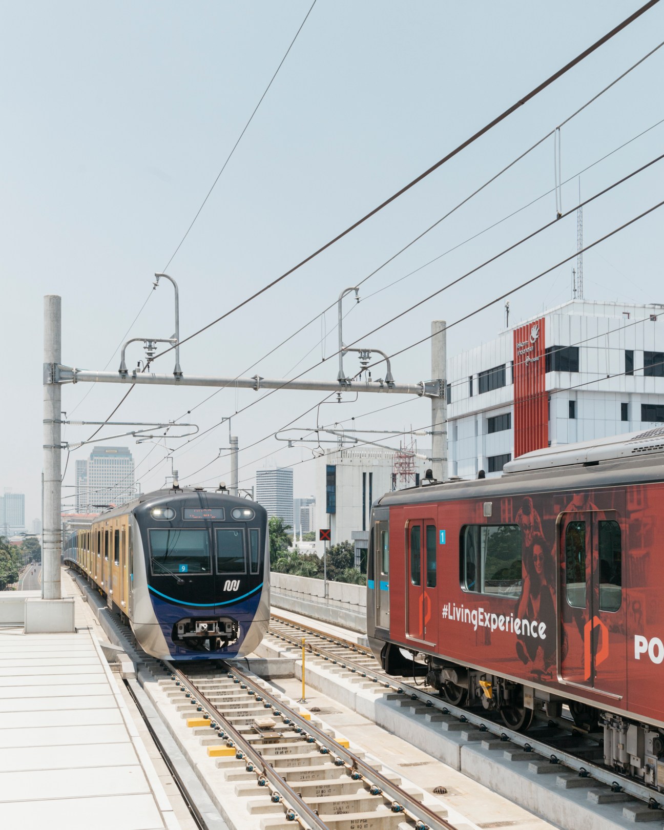 A yellow and blue MRT train pulling into Asean station in Jakarta with overhead power lines and modern buildings in the background.