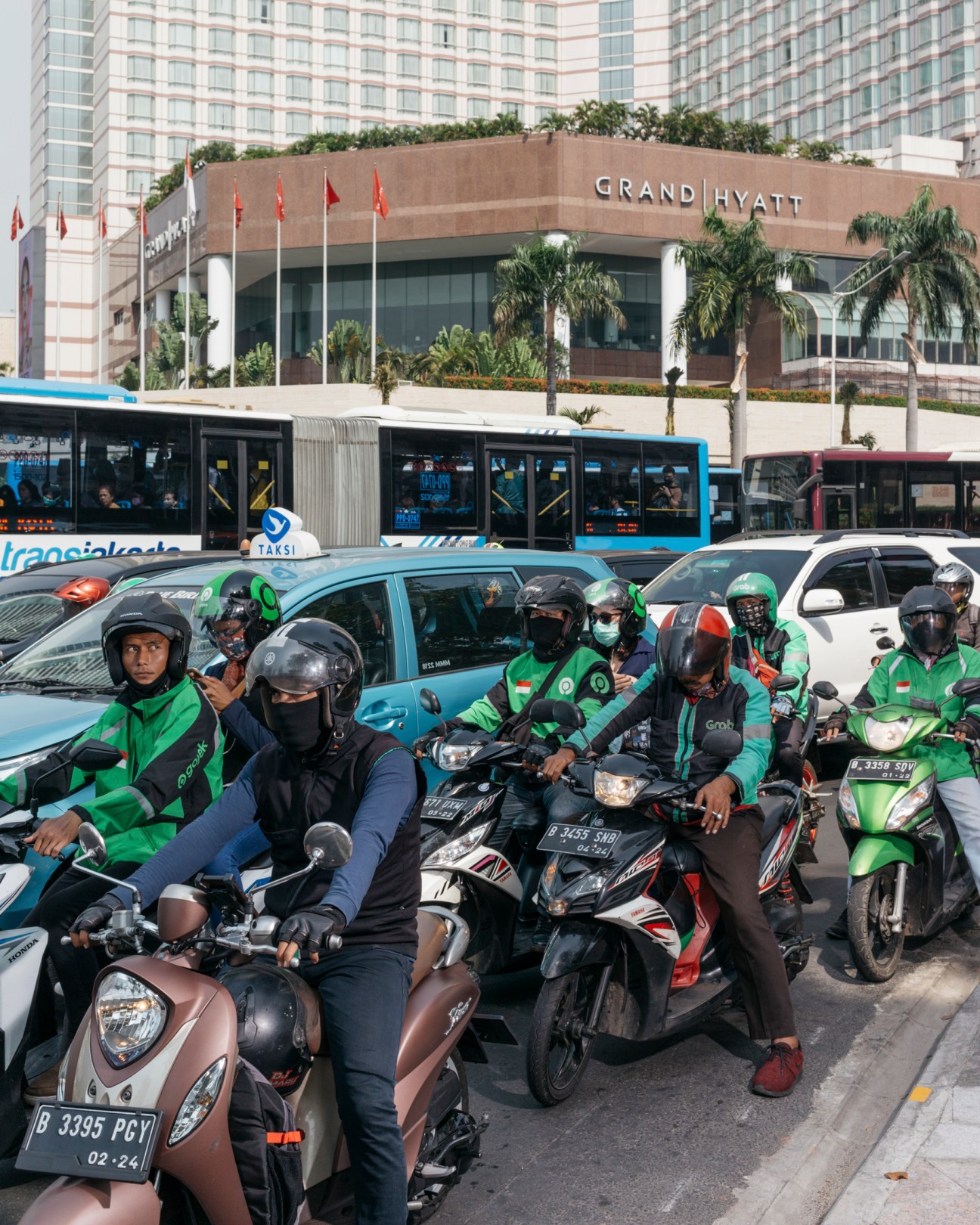 Motorcyclists and taxis congested on a Jakarta street near the Grand Hyatt hotel with red flags and palm trees visible.
