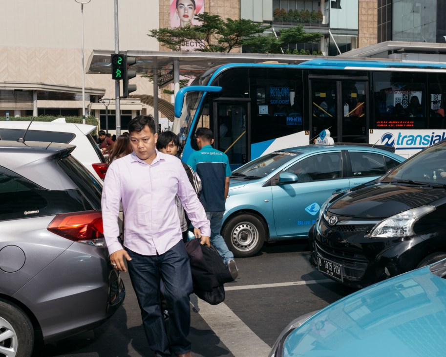 Man in pink shirt crossing street amid parked cars and blue transit bus in Jakarta traffic.