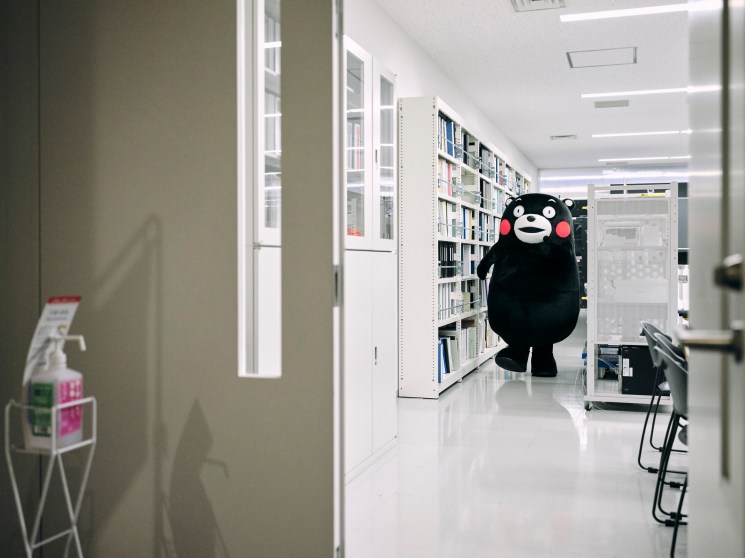 Kumamon, the black bear mascot of Kumamoto Prefecture, stands in a fluorescent-lit office corridor lined with shelves.