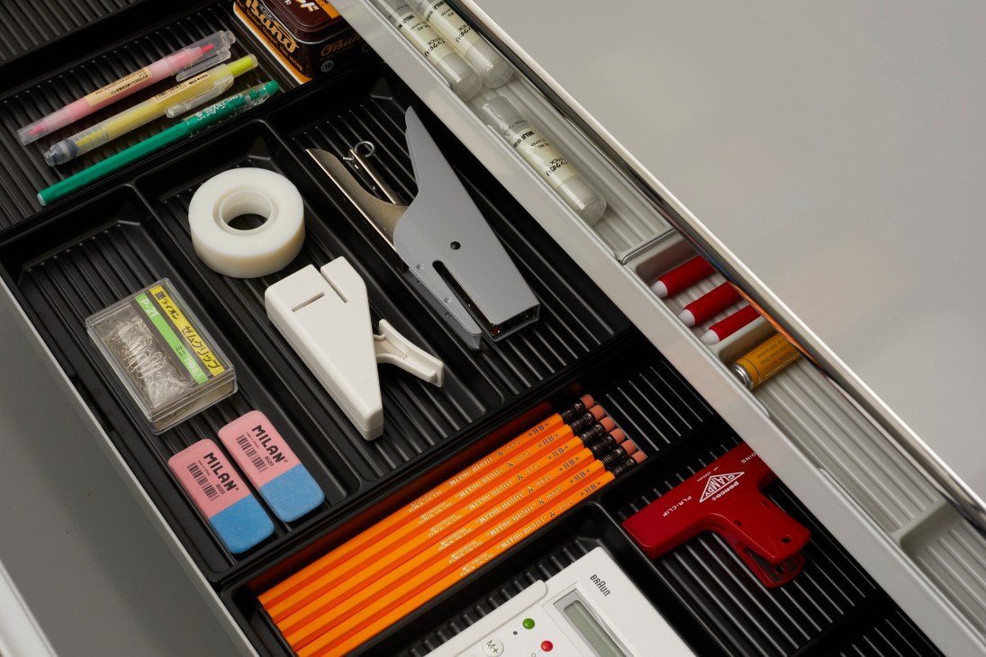 Organized stationery drawer containing colored pencils, tape dispenser, erasers, ruler, calculator, and various writing instruments.