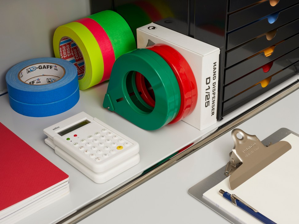 A tape dispenser by Present & Correct alongside colored tape rolls, calculator, and clipboard with pen on a white surface.