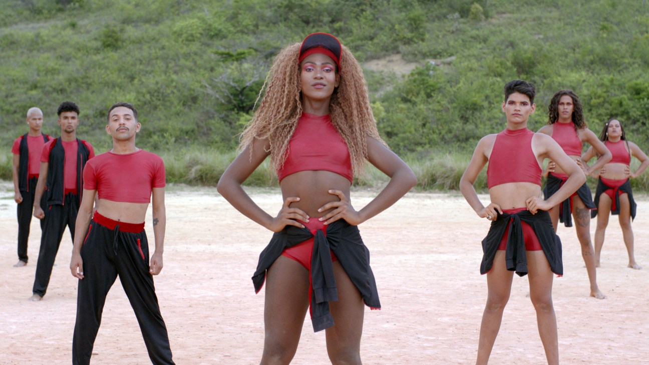Dancers in red and black costumes performing choreography on sandy ground with green vegetation in background