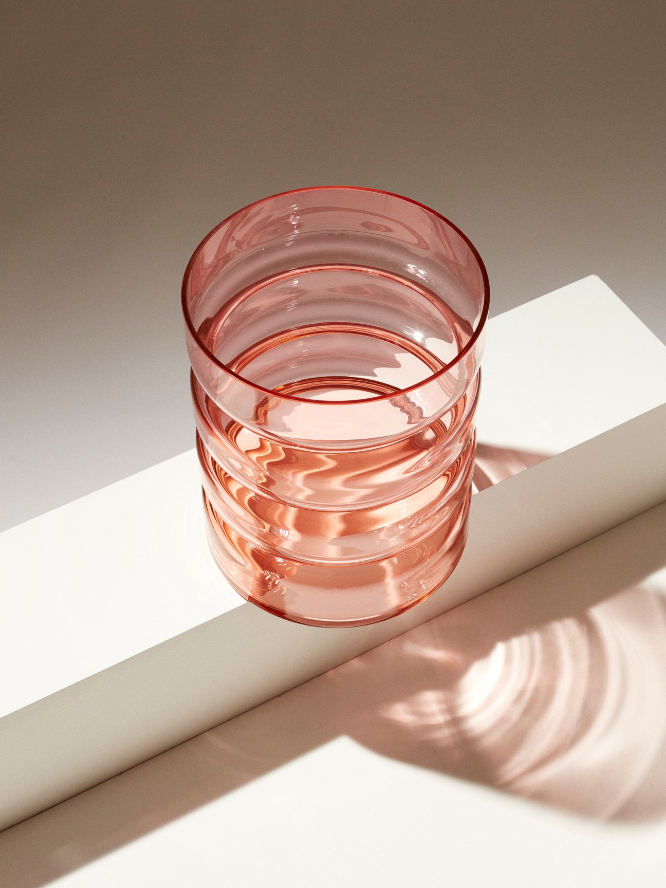 Pink glass tumbler with ribbed design casting shadow on white surface
