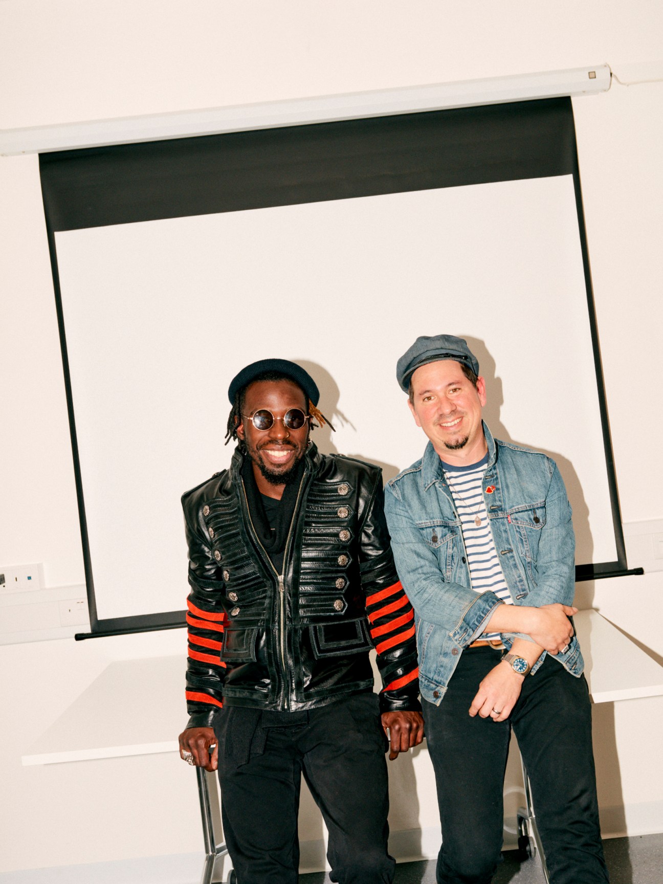 Two musicians sitting together, one wearing a black military-style jacket, the other in denim