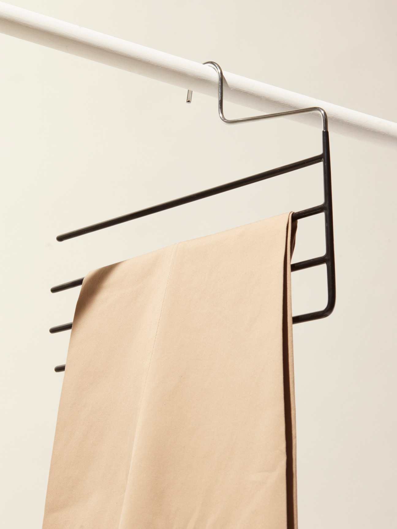 Black metal trouser hanger with multiple horizontal bars hanging from white closet rod with beige trousers