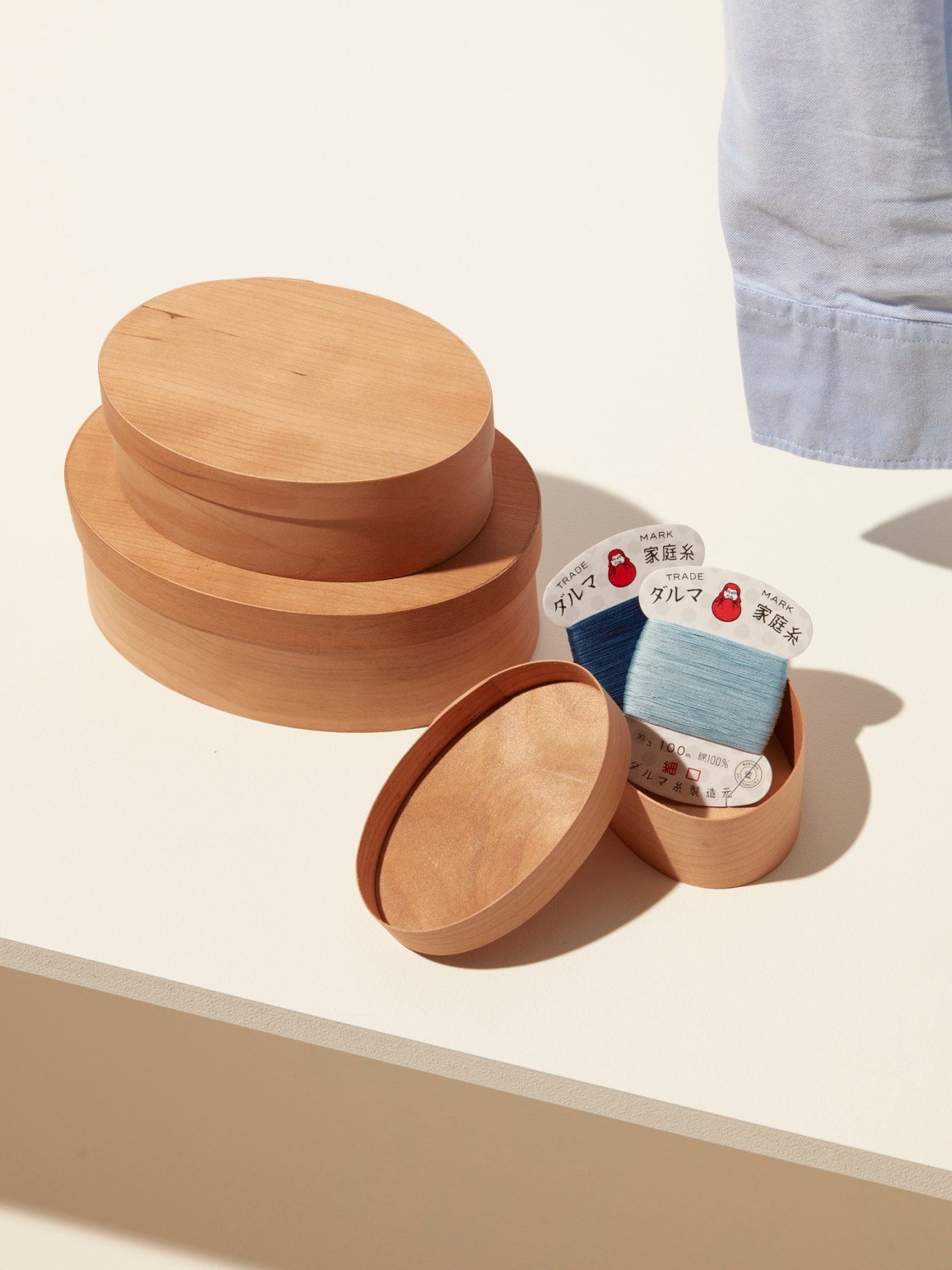 Wooden circular shaker boxes stacked with sewing kit containing blue thread spools on white surface