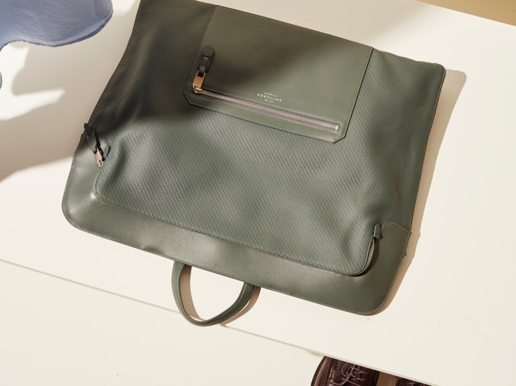 Olive green Smythson leather clothes bag with zipper compartment on white surface next to dress shoes