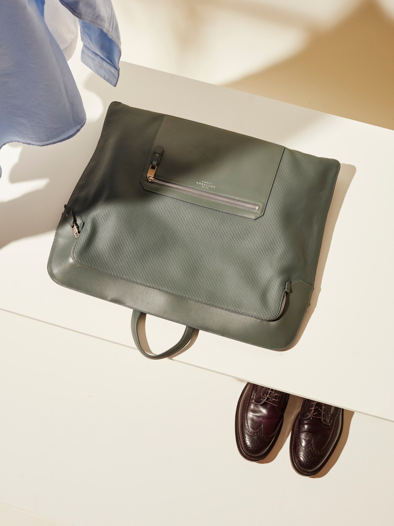 Olive green Smythson leather clothes bag with zipper compartment on white surface next to dress shoes