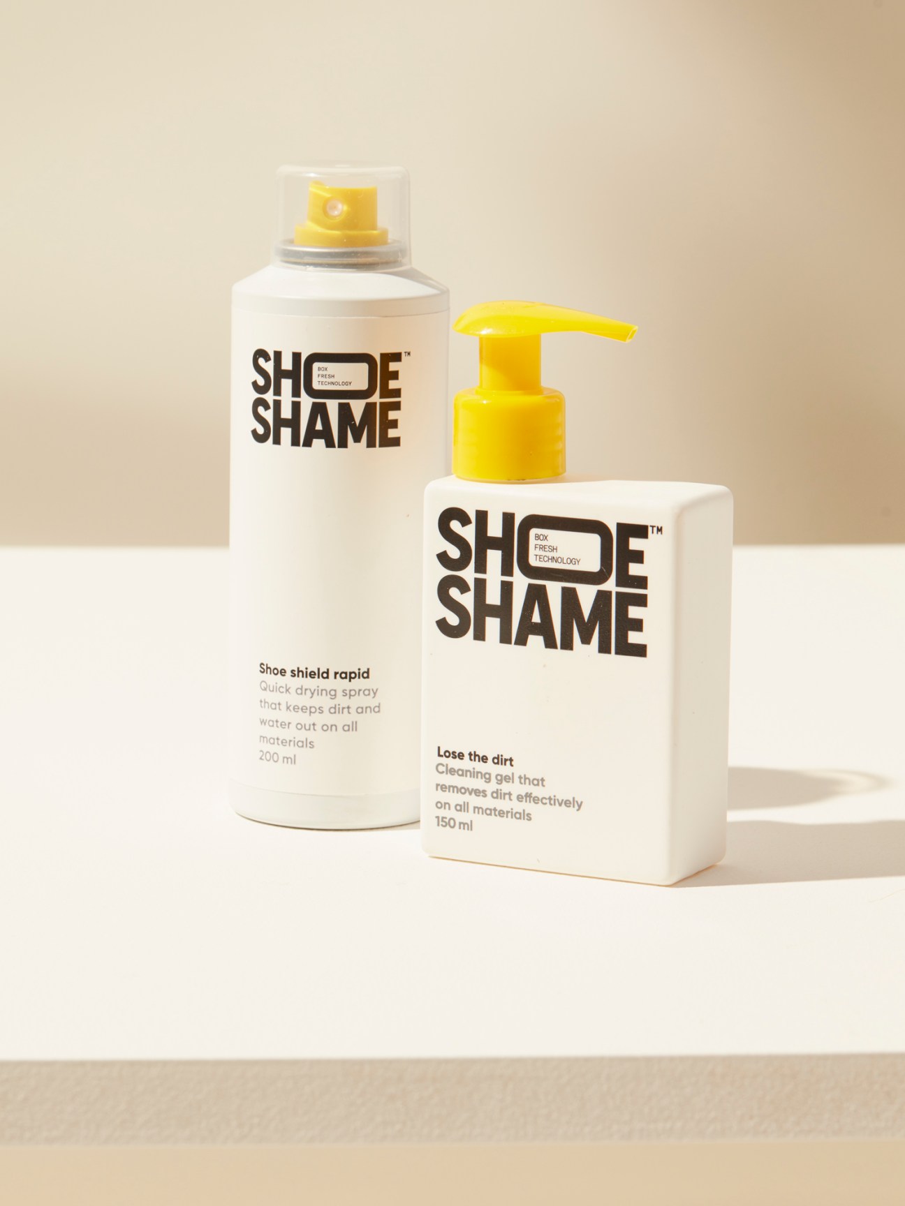 Two white Shoe Shame shoe care products with yellow dispensers on beige background