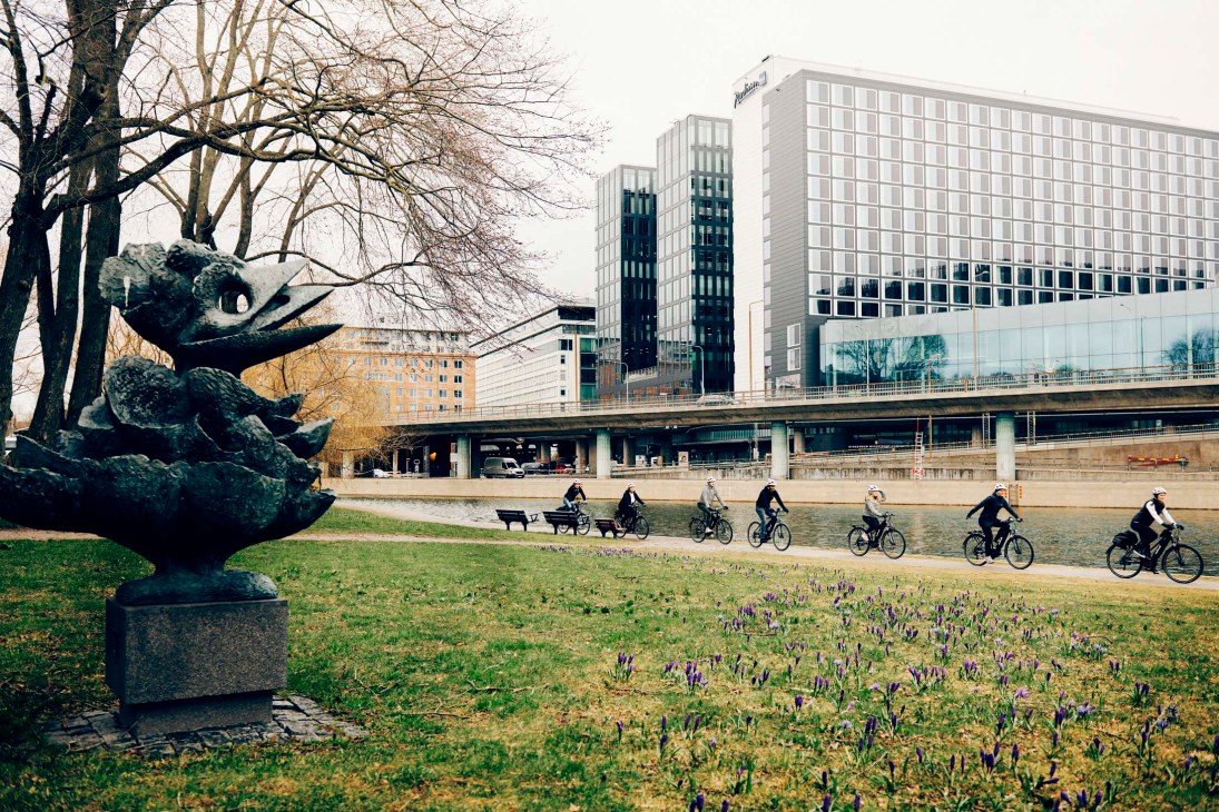 Group of cyclists riding along a waterside path in an urban park with modern buildings in the background.