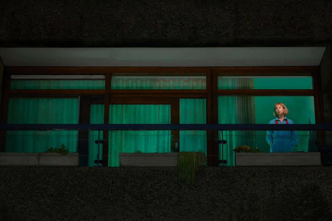 A person in a blue shirt stands at a window with green curtains during lockdown isolation.