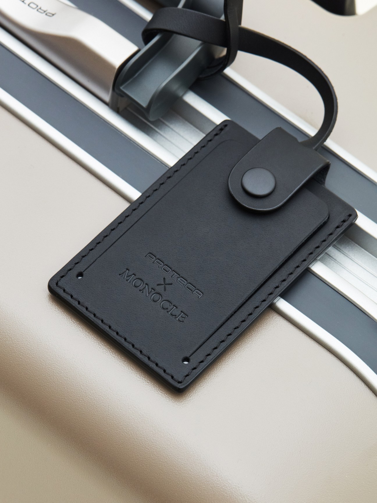 Black leather luggage tag with Proteca x Monocle branding attached to light-colored suitcase handle