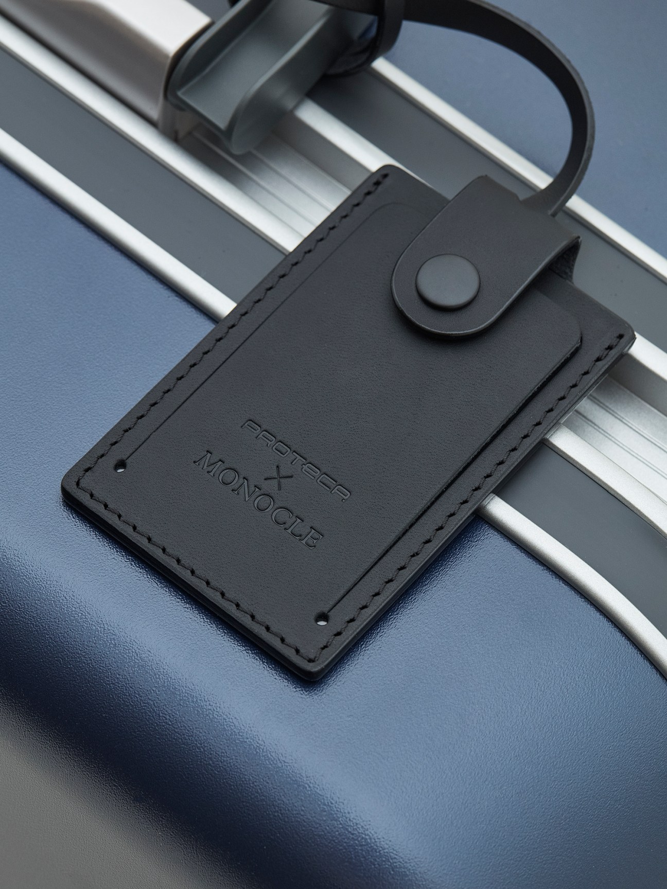 Black leather luggage pouch with Proteca x Monocle branding attached to navy suitcase handle.