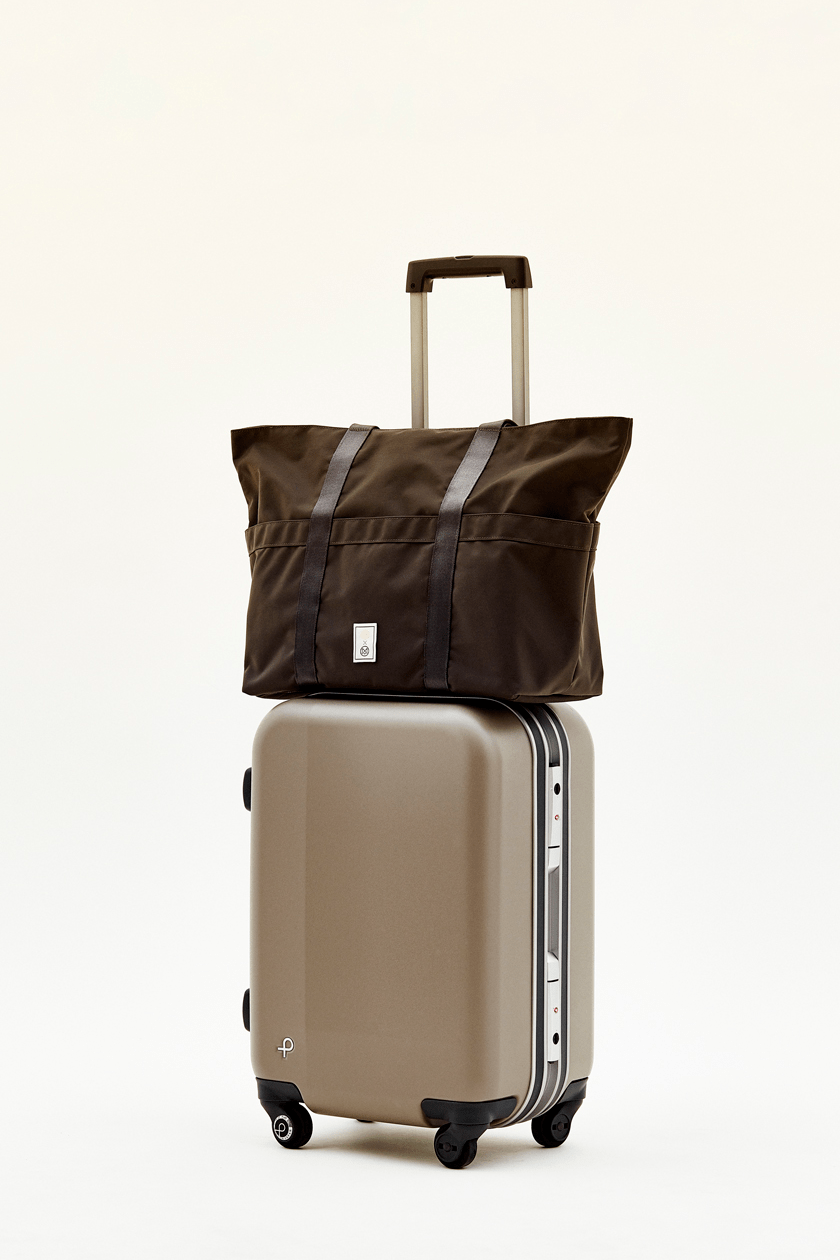 Brown trolly tote bag by Monocle positioned on top of a beige Proteca carry-on suitcase with retractable handle.
