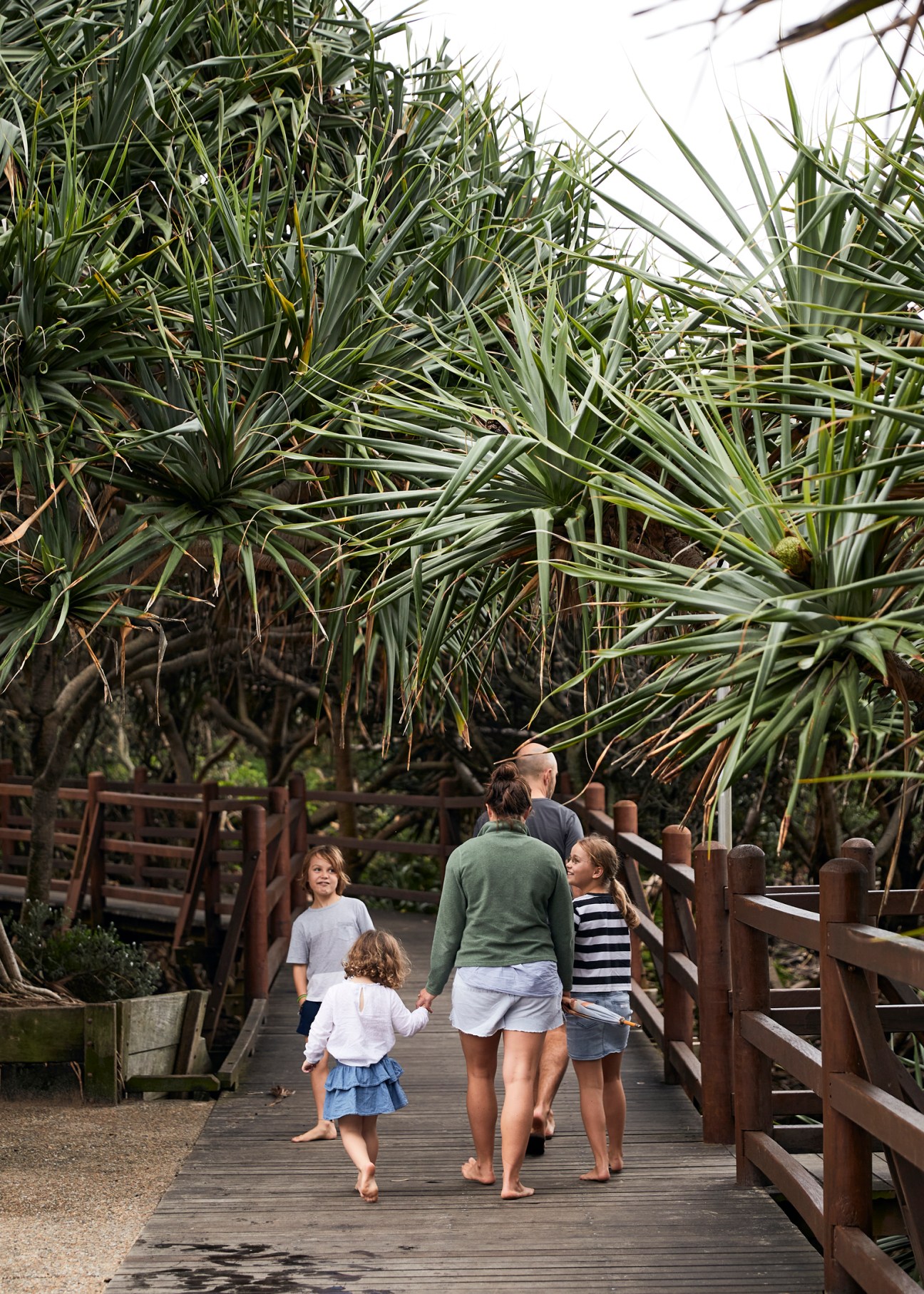 Family of five walking on wooden boardwalk surrounded by pandanus palms and coastal vegetation