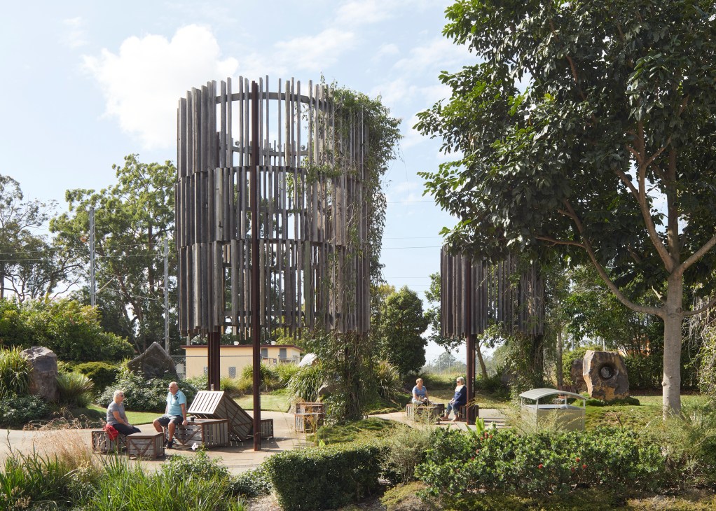 Cylindrical vertical wooden structures in landscaped public space with people sitting on benches surrounded by trees