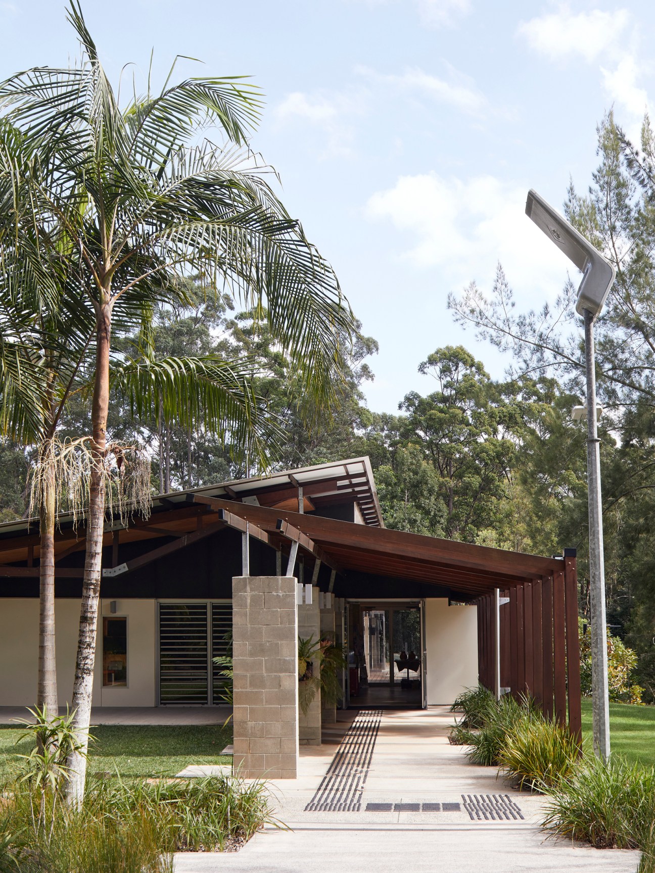 Modern single-story building with timber overhang, concrete block pillar, and solar street lamp amid tropical vegetation