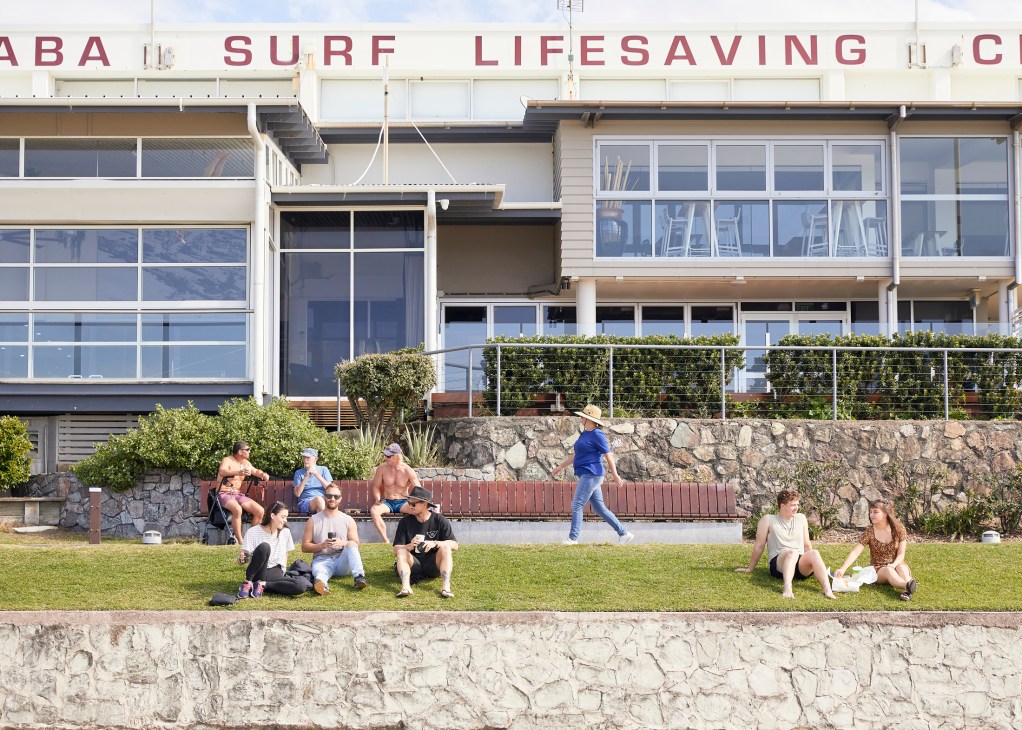 People relaxing on grass in front of a surf lifesaving club building with large windows