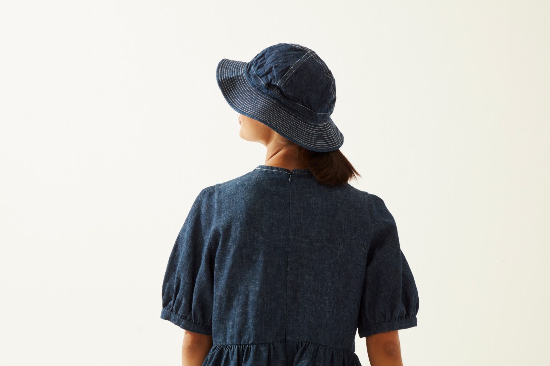 Woman wearing dark denim dress with puffed sleeves and wide-brimmed denim hat, photographed from behind.