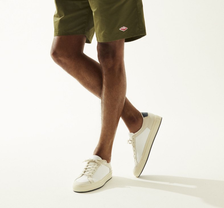 Man wearing olive shorts and cream lace-up trainers against white background.