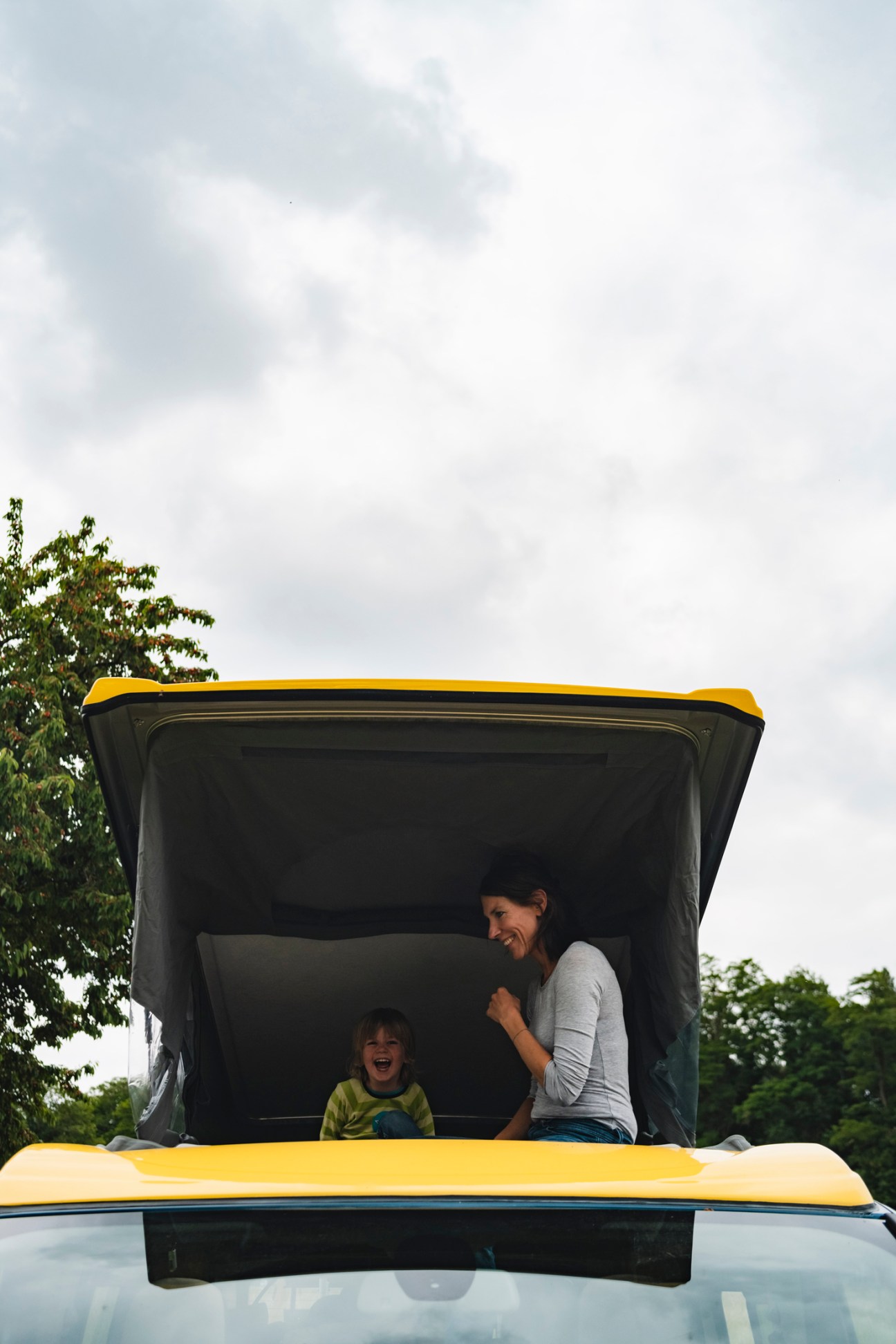 Woman and child sitting in the open rear of a yellow and gray camper van with pop-up roof extended