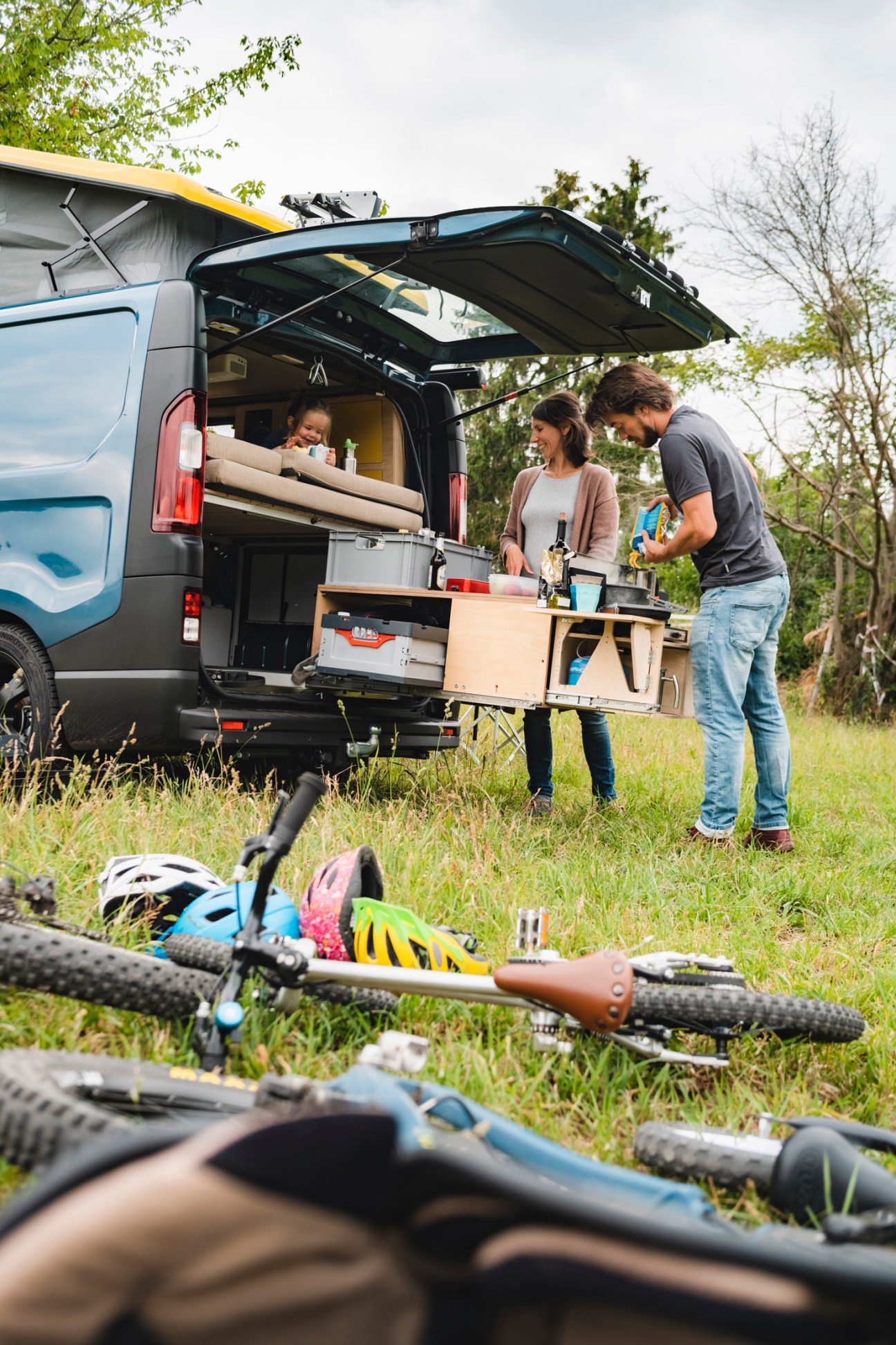 Family with camper van kitchen setup outdoors, bicycles in foreground on grass